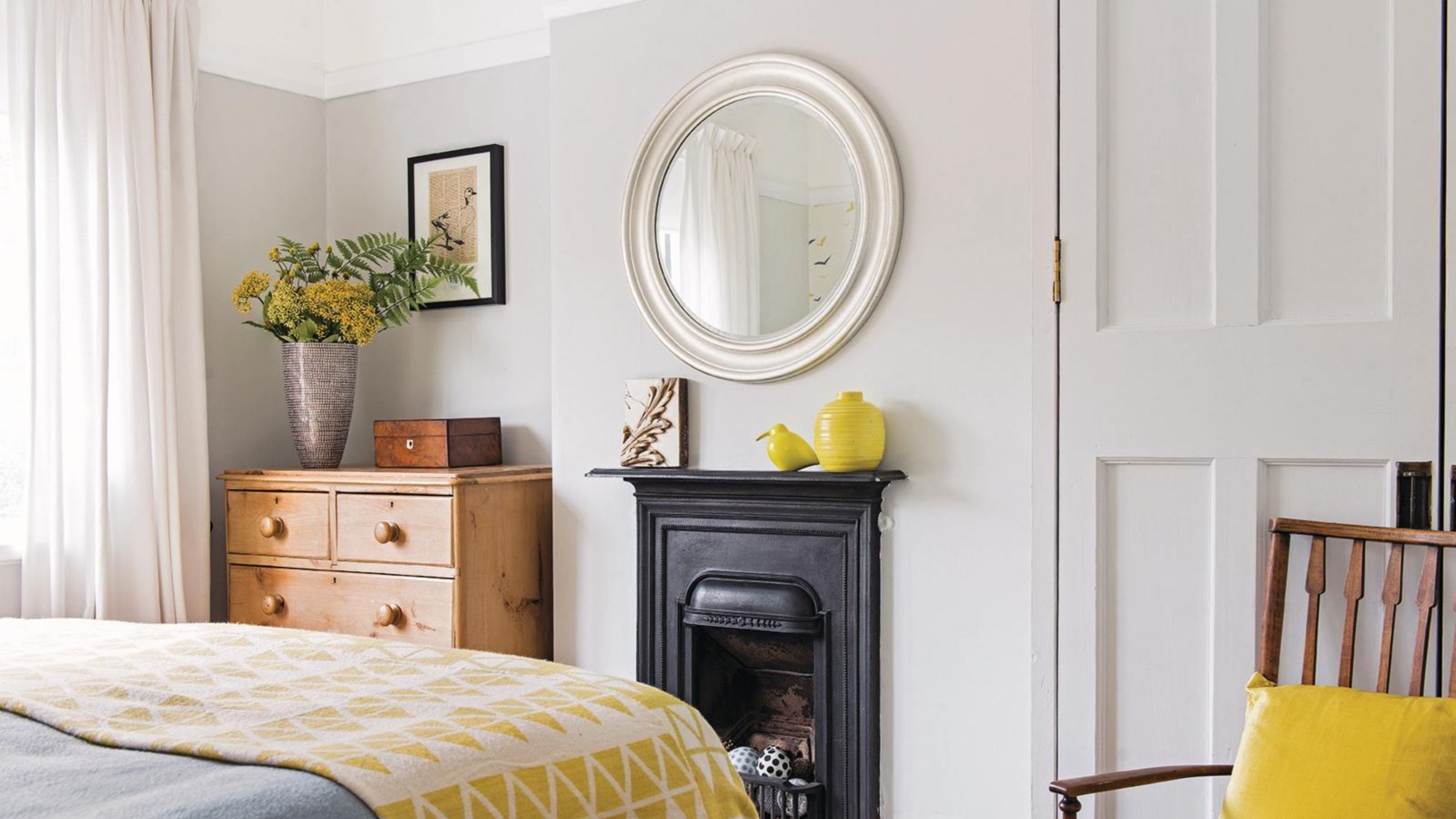 A grey bedroom with a wooden set of drawers, and a small black fireplace with a cream mirror hanging above. 