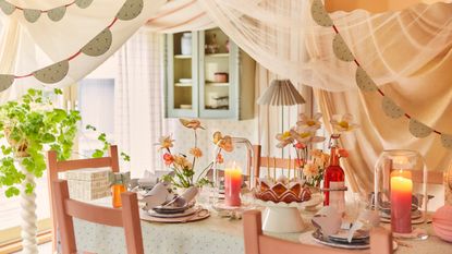 Image of a pastel dining table area with sheer fabric draping from the ceiling and garland made of wallpaper. The table is set for a spring dinner party and there are flowers and bows made of wallpaper on the table as well.