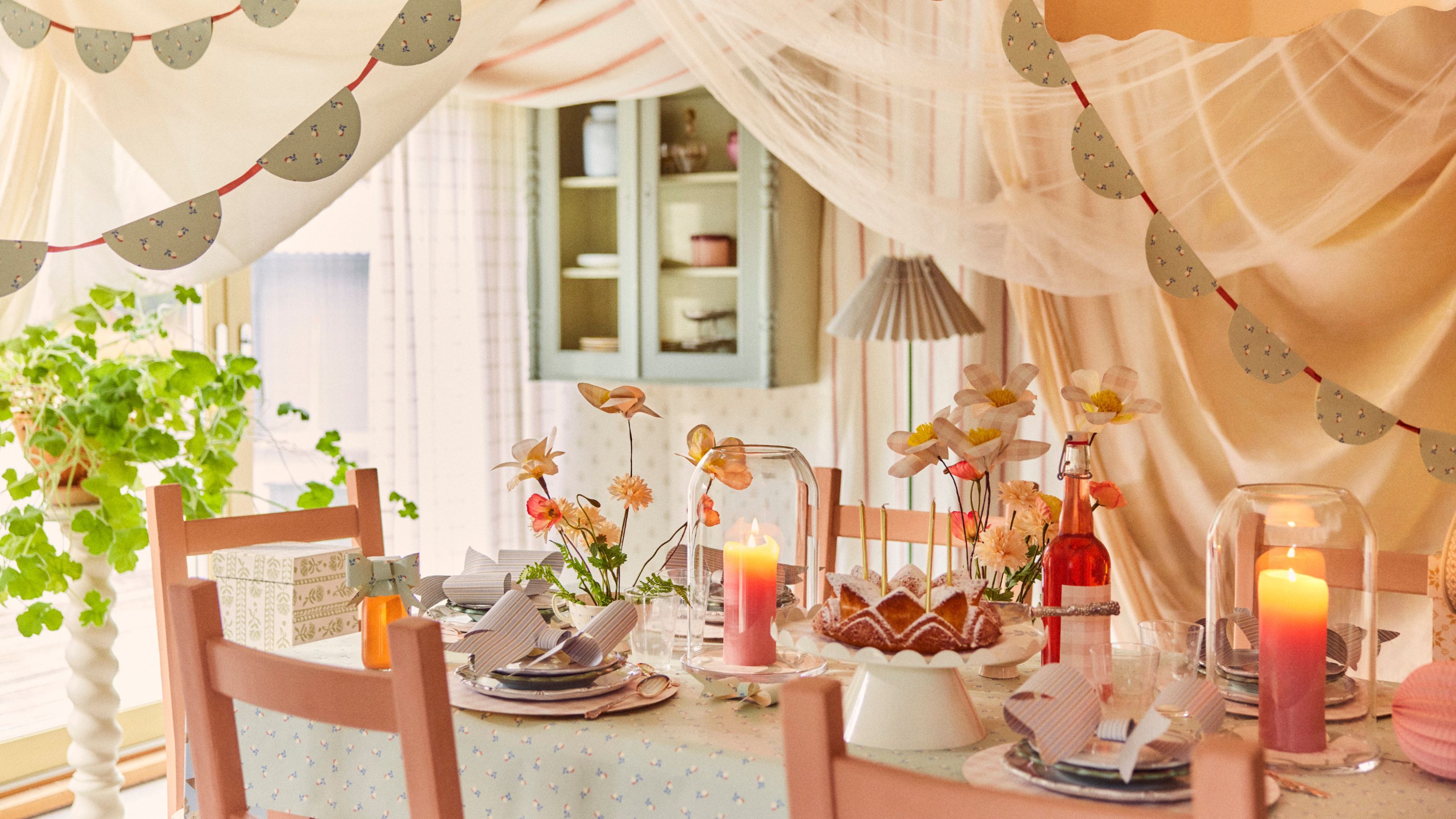 Image of a pastel dining table area with sheer fabric draping from the ceiling and garland made of wallpaper. The table is set for a spring dinner party and there are flowers and bows made of wallpaper on the table as well.