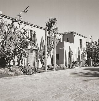 Black and white image of The Main House at Lotusland, Montecito, California, 1919, stone slab driveway, cactus plants of various sizes decorate the front the house, slate roof, windows, clear sky
