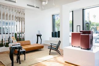 The brightly lit room of a modernly furnished shop features a silver, reflective wall, mid-century furniture and a podium with red suitcases standing on it.