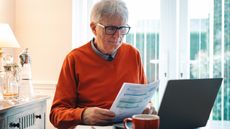 An older man looks over pension paperwork while sitting in front of his laptop in the kitchen.