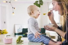 a medium shot of a blonde-haired mum baby-weaning her child with a spoon in the kitchen