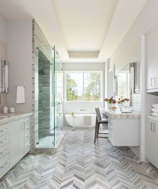 Large bathroom with recessed white ceiling, herringbone gray tile floor in marble finish, looking on to small white bathtub, shower with glass door, off-white cabinets with marble countertop, seating area, tiled walls
