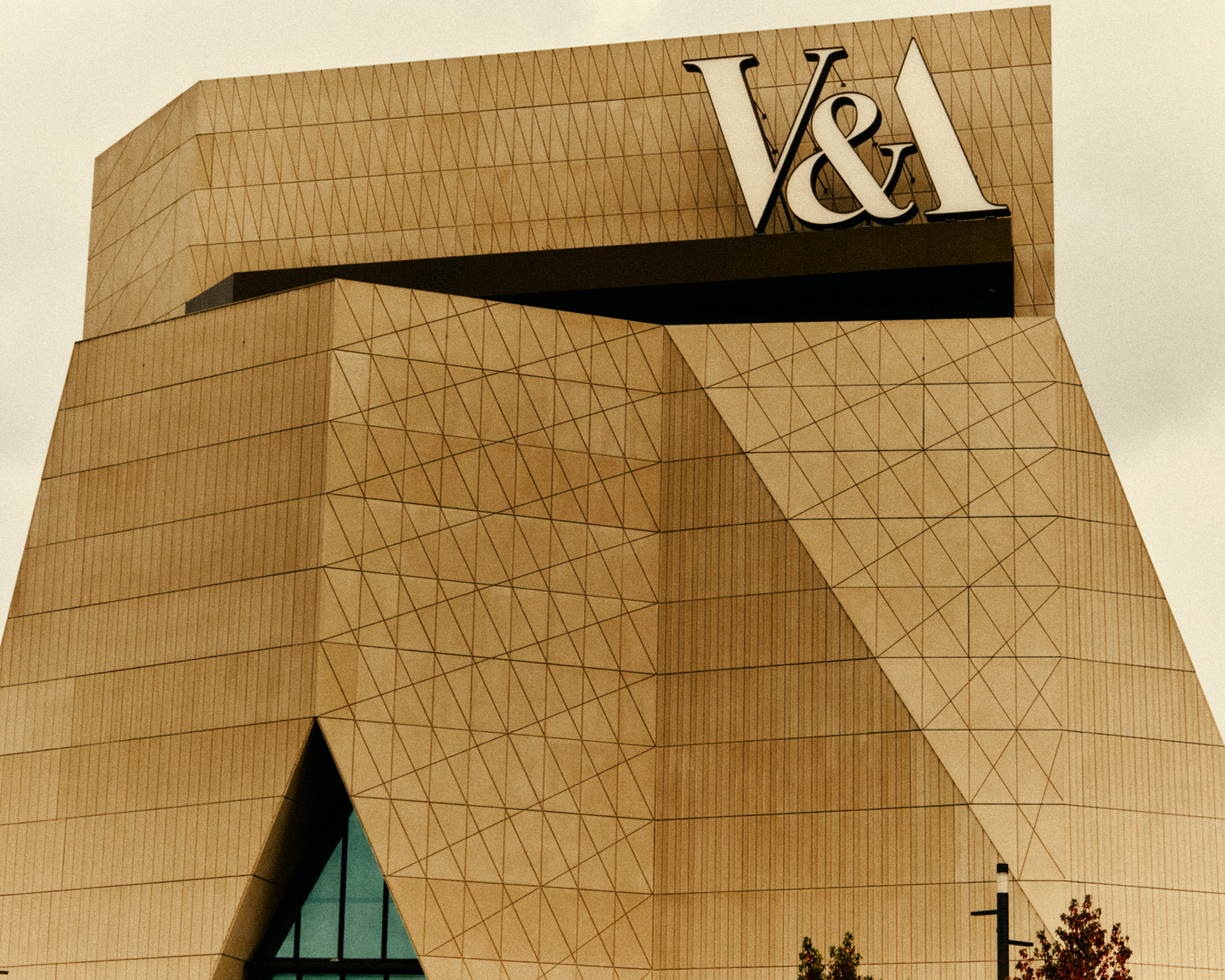 Exterior view of a geometrically shaped museum facade with multiple cement slabs combined into an angular shape, with a white sign that reads, in caps, "V&amp;amp;A".