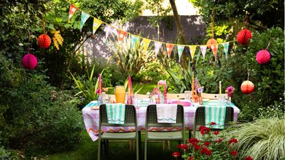 Pinterest summer decor trends are so stylish. Here is a backyard space with a rectangular table with pink gingham cloth, glasses, colorful candles and floral placemats on it, green and wooden chairs around it, multicolored gingham bunting above it, and trees and fencing