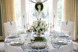 A blue and white formal dinner setting set at a round table. There is a white floral arrangement in the center and three white chairs