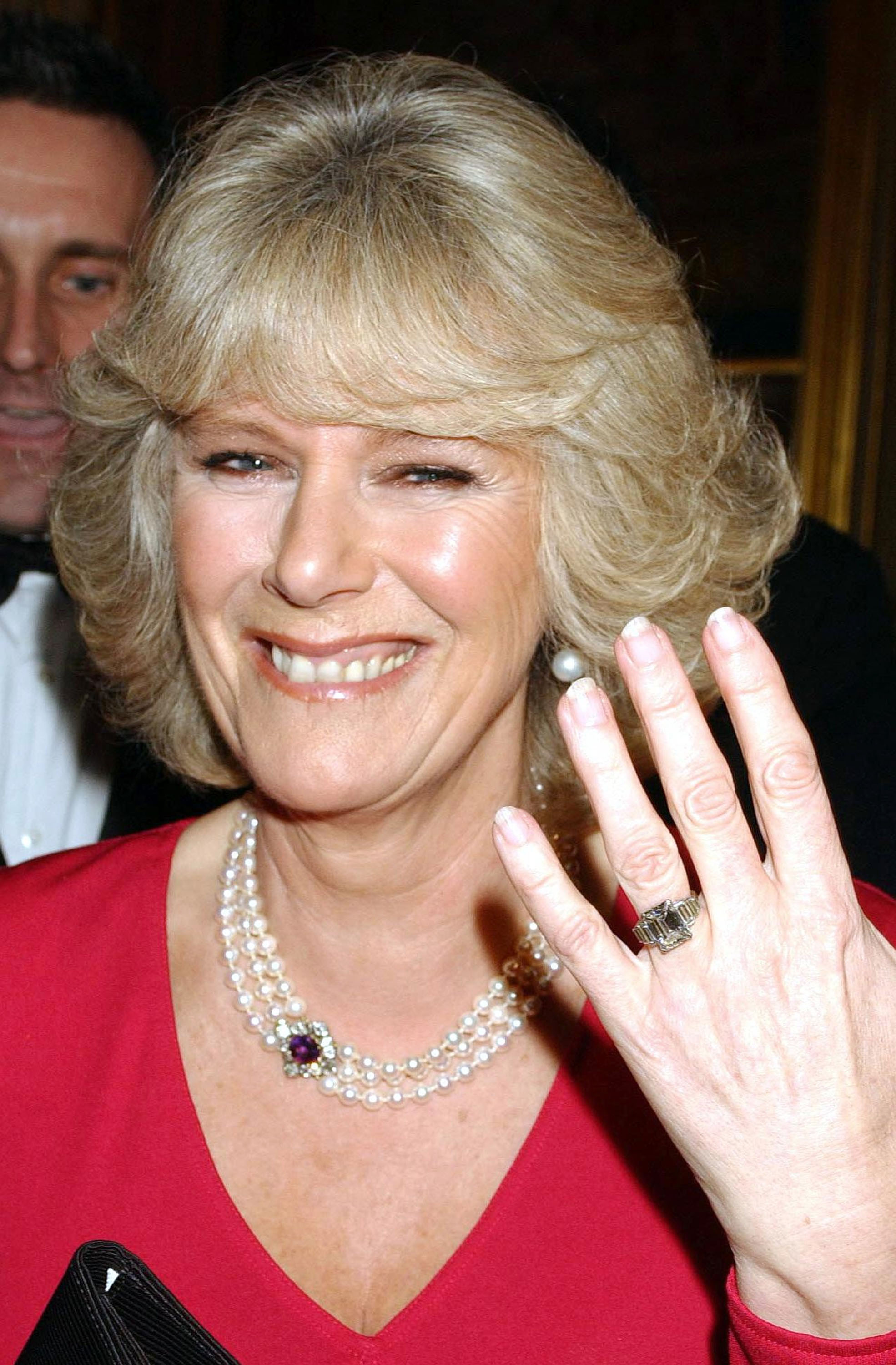 Queen Camilla wearing a red dress and pearls holding her hand up to the camera to show her engagement ring