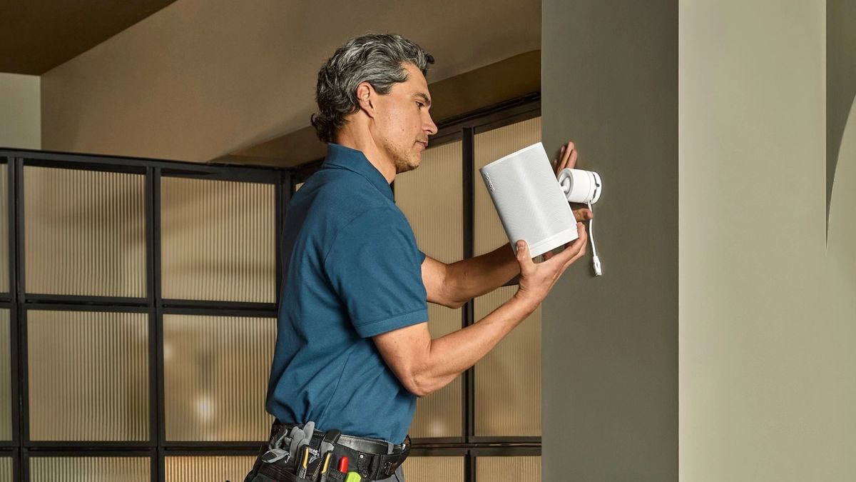 A man in a blue shirt is attaching the speaker to a wall. The speaker is white. 