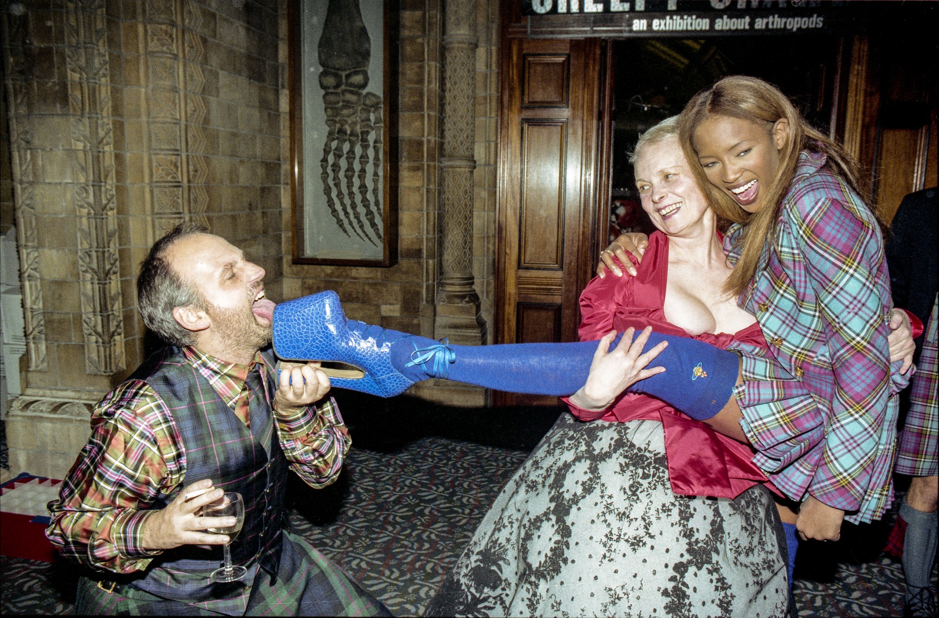 Colour photo of hairstylist Sam McKnight kneeling and licking a blue platform boot held up by Naomi Campbell, who laughs as Vivienne Westwood holds her leg at a party in the Natural History Museum.