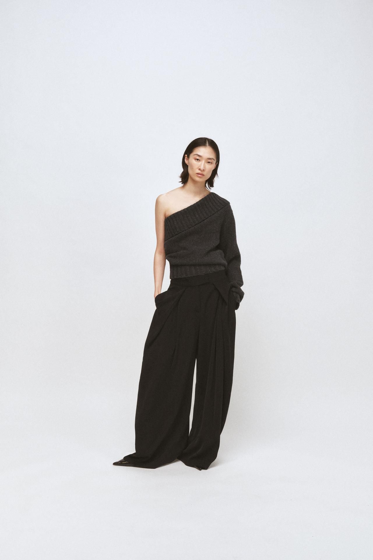 model wears one shoulder longsleeve sweater in gray and wide-leg slouchy black trousers with a pointed black heel.