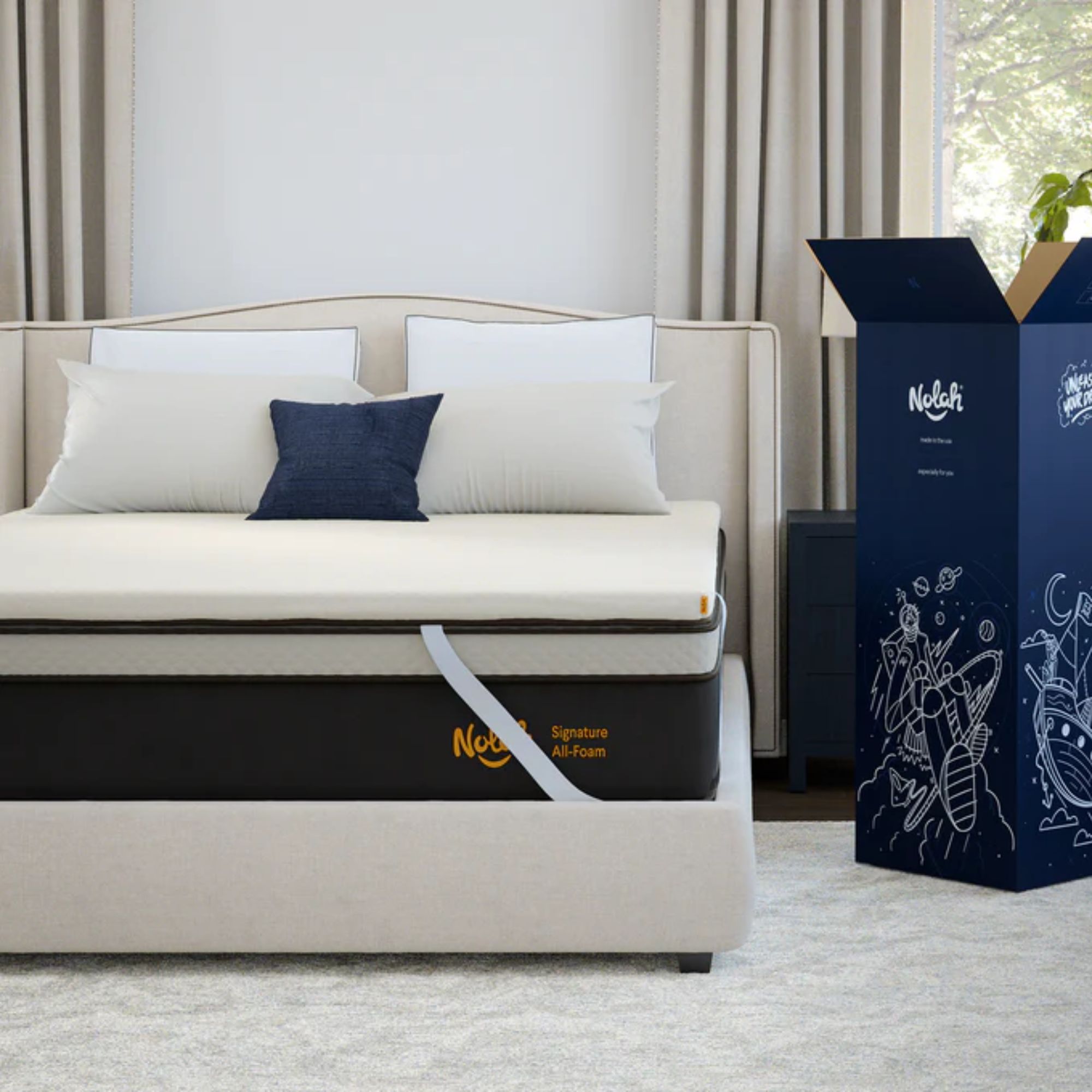 Nolah mattress topper on a white upholstered bed beside a large blue packaging box, on top of a cream rug. Behind is a white wall, and to either side of the bed are cream curtains, with a large window seen to the right.