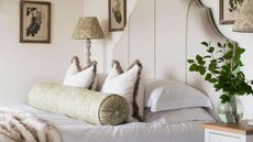 A bed with a tall beige headboard, scatter pillows and a plant on the side table