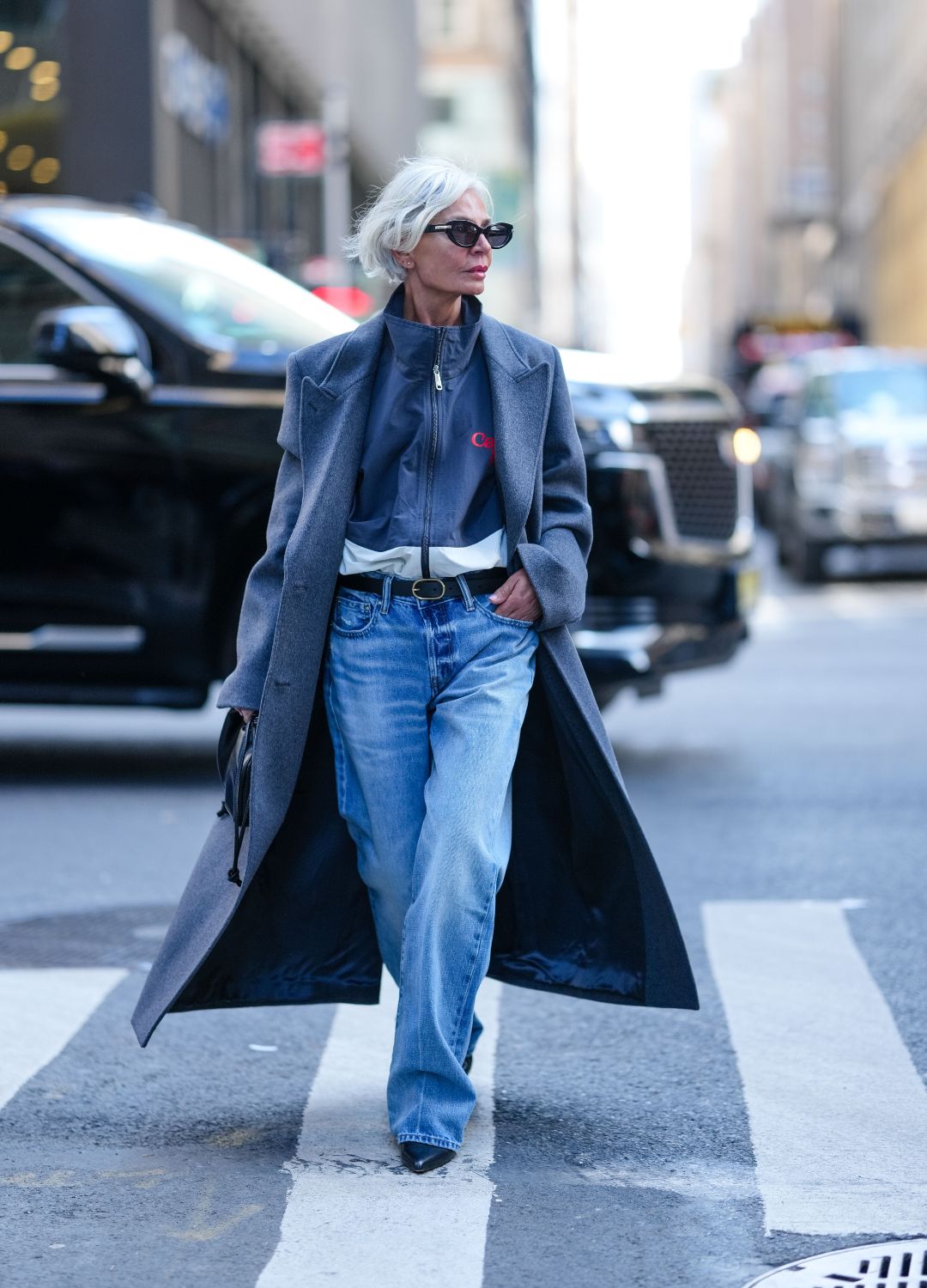Grece Ghanem wears black sunglasses, dark navy blue white zipped high neck jacket, dark blue double breasted wool coat, shiny black Calvin Klien drawstring leather bag, black leather belt, light blue washed loose denim jean pants, shiny black pointed toe kitten heels leather shoes, outside Calvin Klein, during the New York Fashion week Fall/Winter 2025 on February 7, 2025 in New York, New York