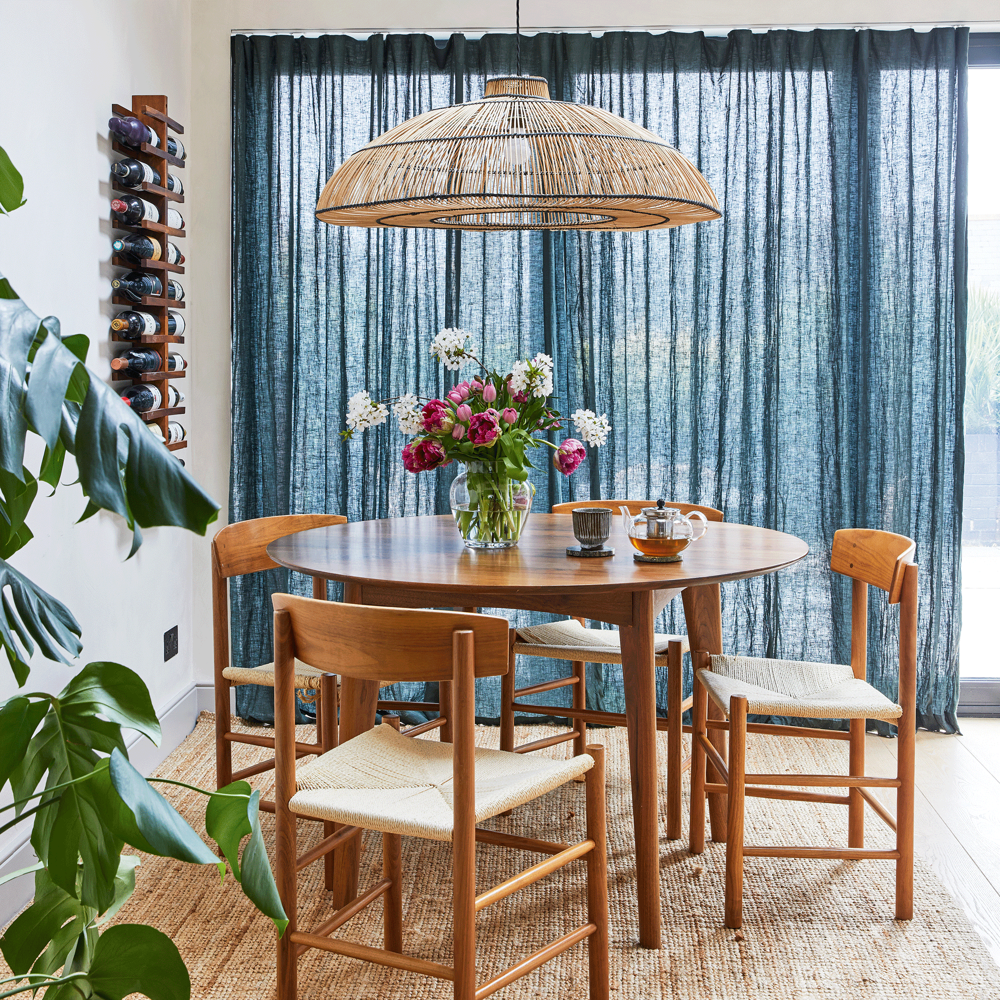 a dining area in an open plan room, a mid-century round dining table and chairs with a rattan pendant shade, and a wall-hung wine rack