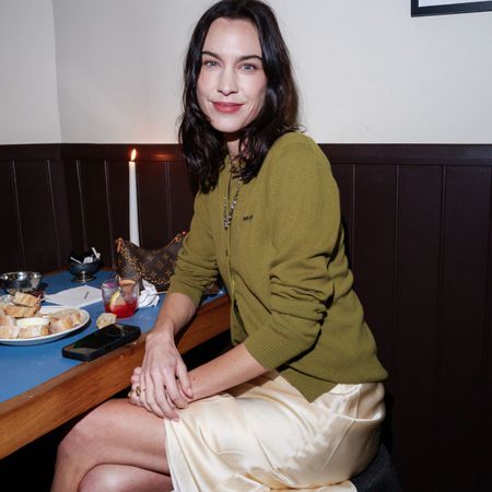 An image of Alexa Chung wearing a Reformation slip dress, grey coat, olive cardigan and gold heels. 