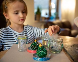 Children making Christmas snow globe decorations with glass jars, glitter and pine cones