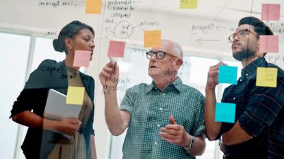 A gray-haired, balding man explains a formula to two younger workers.