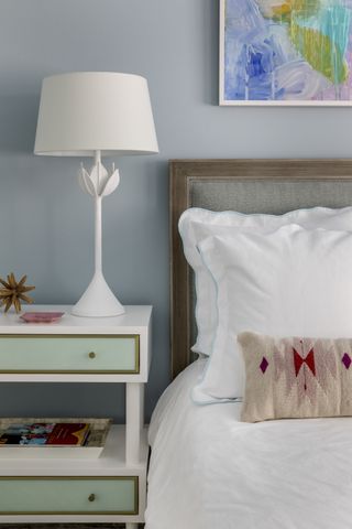 pastel blue bedroom with green and white side table, bright multicoloured artwork, white lamp, white bedding, pink and stone cushion