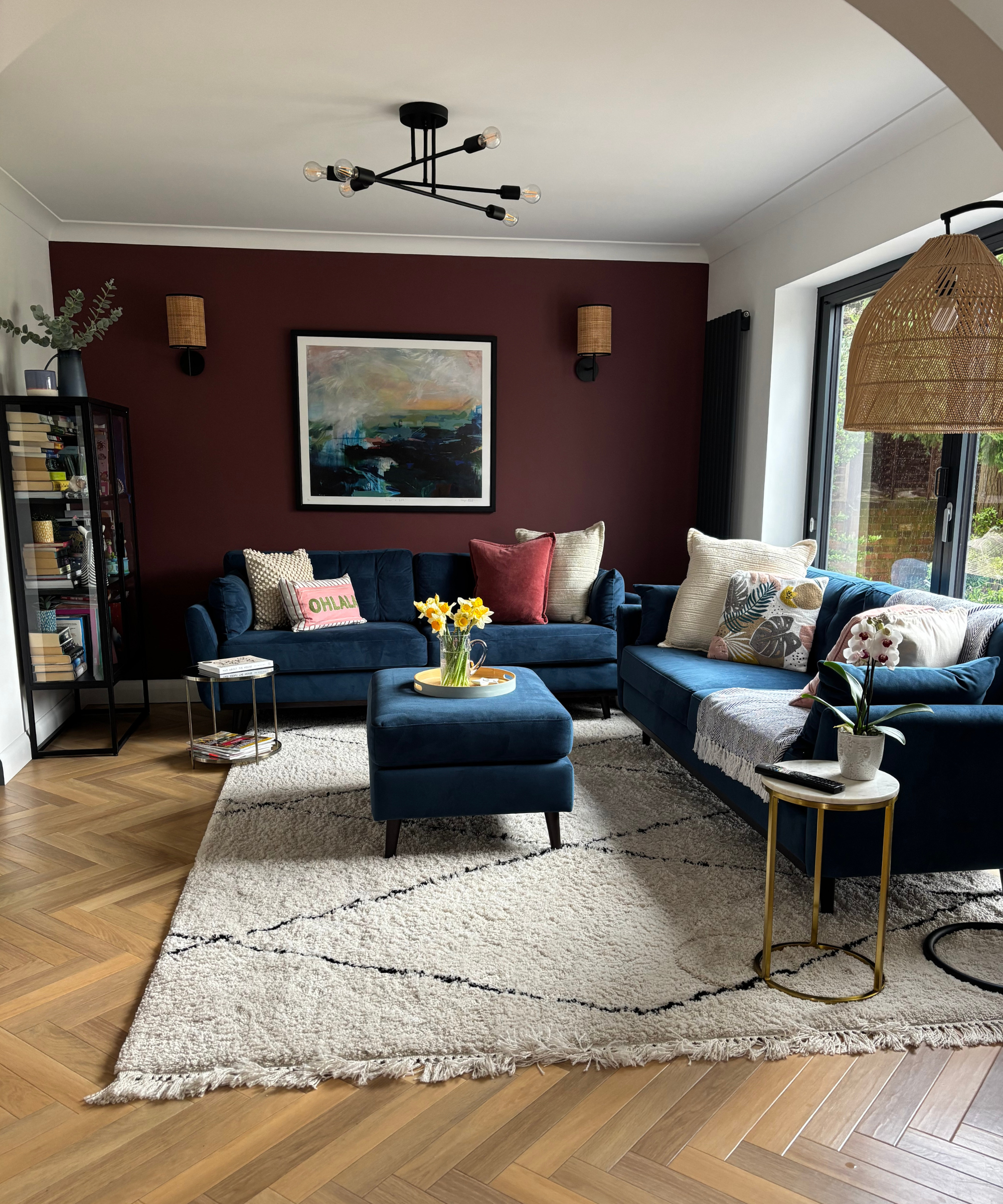 Laura's living room with a burgandy feature wall, blue sofas, wooden flooring, and lamp in the right corner