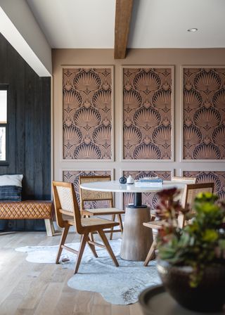 A dining room with a round table, cane chairs, and wall panels filled with art deco style wallpaper