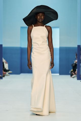 A female model wearing a long pink sleeveless dress and a large black hat walking down a runway.