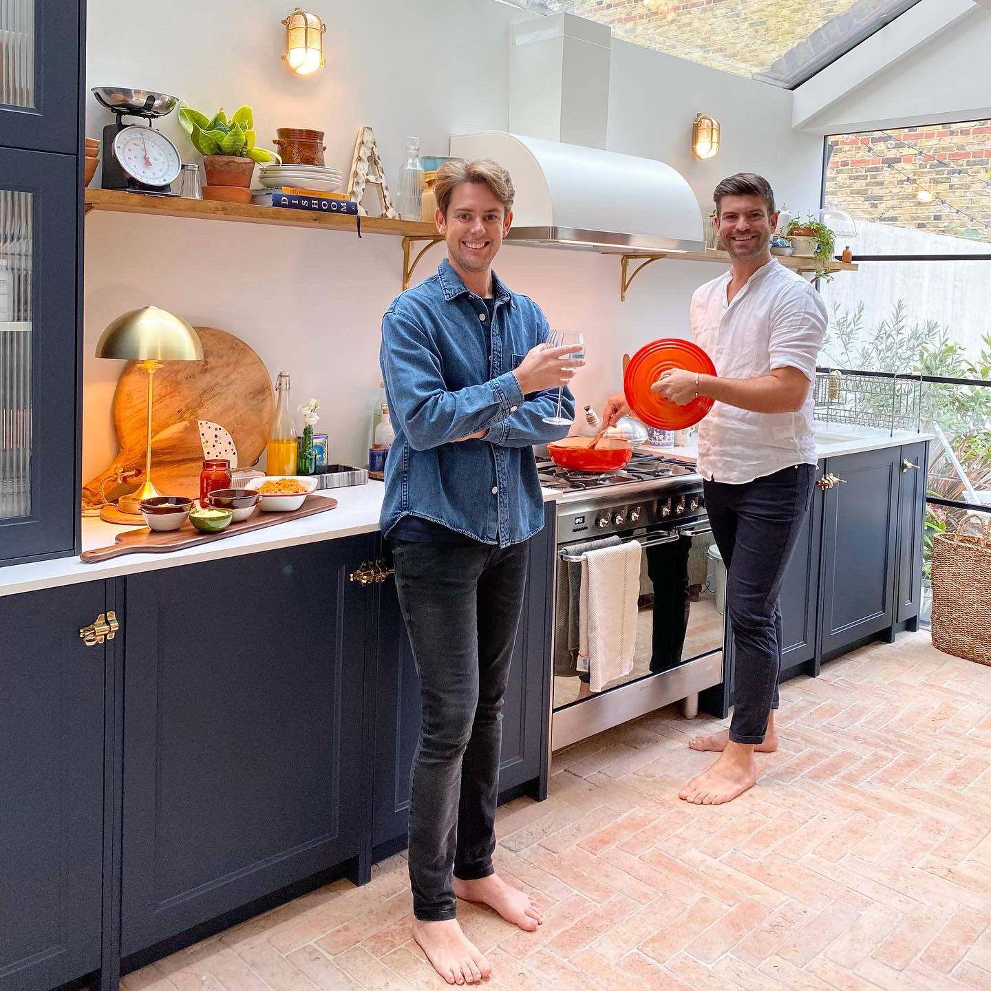 Two men standing in a navy kitchen with glass roof and terracotta floor tiles, cooking with an orange cast iron pan