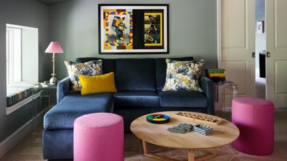 Image of a muted moss green living room with a navy blue, velvet small sectional sofa. There are two small, round ottomans in a hot pink shade and a small pink lamp on the side table next to the sofa. There is a large round wooden coffee table with games on it.