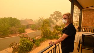 A woman in shown stood on her balcony looking out at the distance. The sky is tinged orange/yellow and she is wearing a respirator mask.