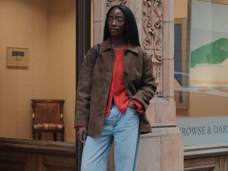 London woman wears a red cardigan with a brown suede jacket, light wash baggy jeans, and loafers.