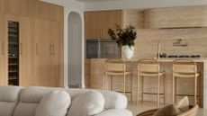 A minimalist kitchen and living room with a boucle style white couch, woven kitchen stools, a white vase of stems, and a beige stone backsplash and island