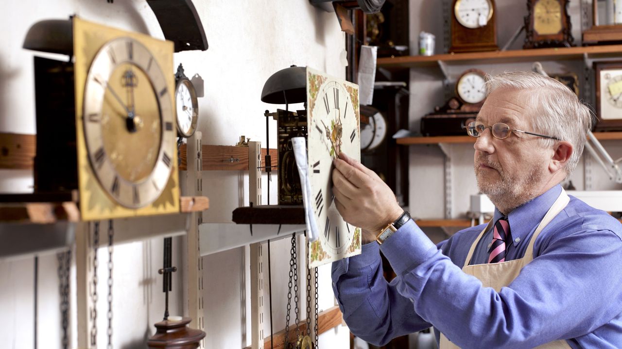 An older man adjusts the time on a clock in his clock repair shop.