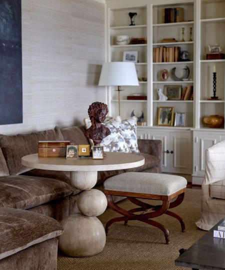 Living room with grasscloth walls, velvet brown sofa, marble round table and antique stool with bookshelves with antiques in background