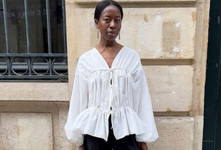 Influencer stands in front of Parisian brick wall wearing white frilly blouse and tailored black pants.