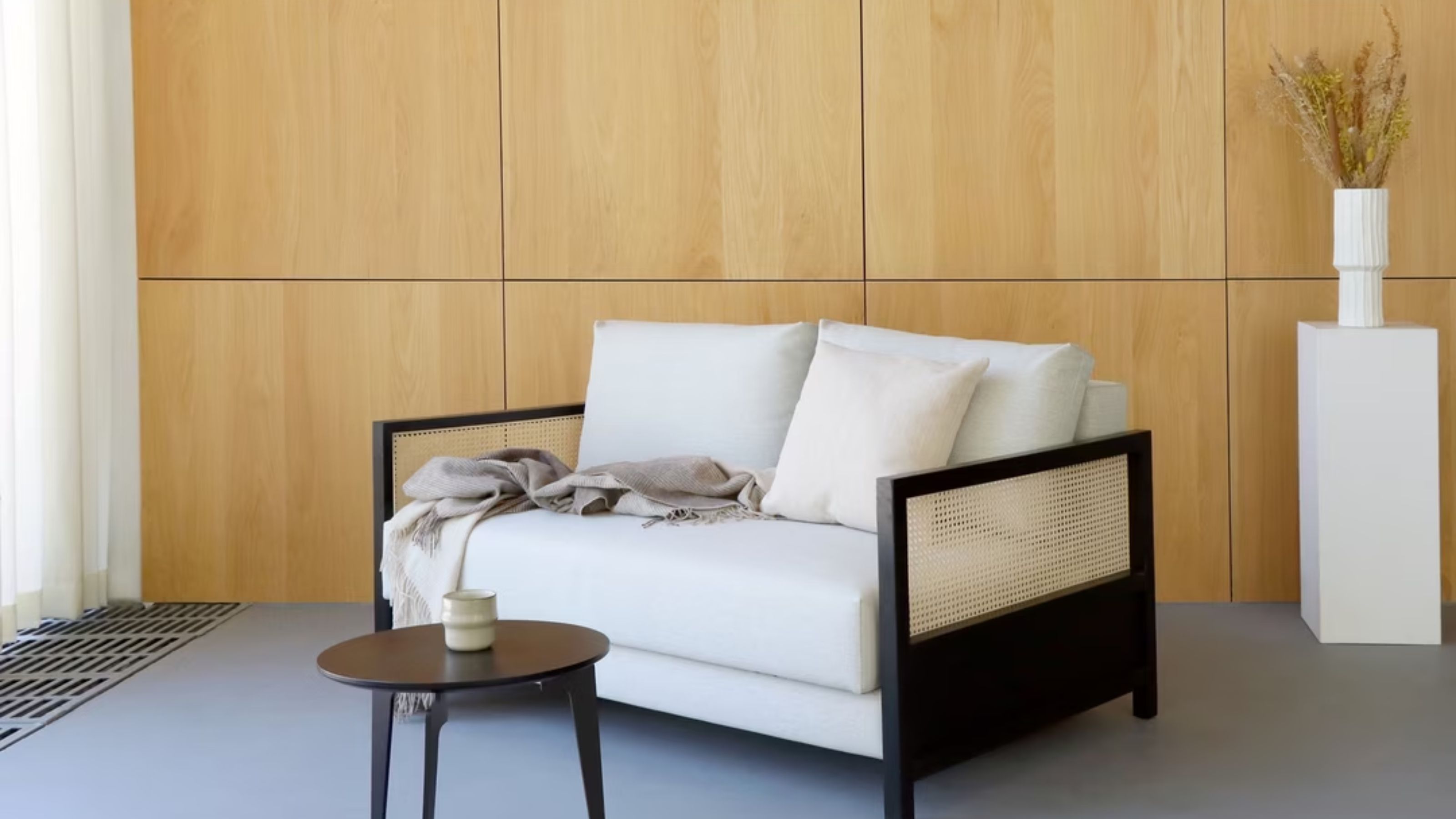 A scandi style sofa bed that had been folding in with a coffee table in front of it as well as a vase and plants on a plant stand in the background of a home 
