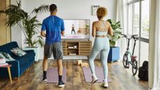 Woman and man exercise in front of a screen, on which a trainer can be seen. The man wears grey shorts and a blue shirt, the woman is in a grey two-piece. 