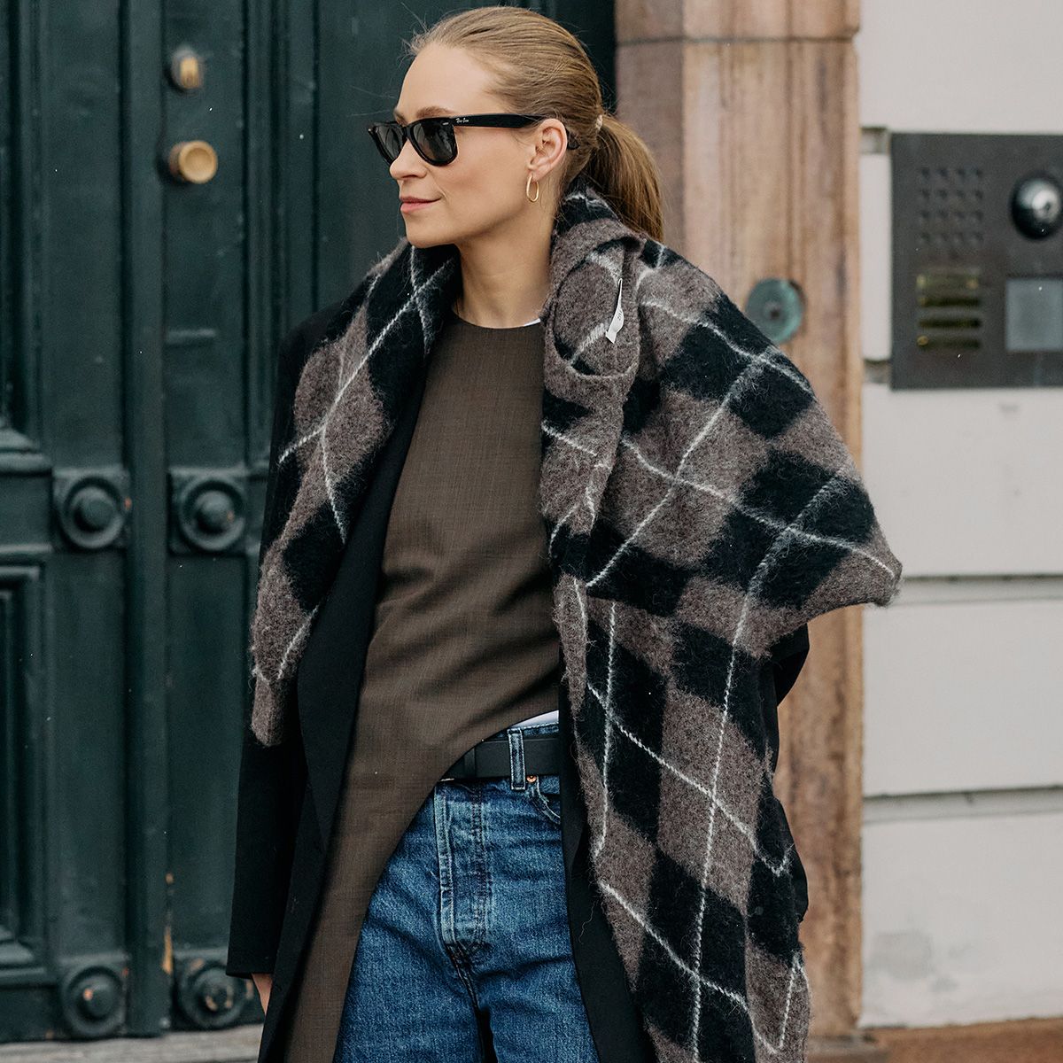 A woman wears a plaid sweater, brown top, jeans, and a black coat.