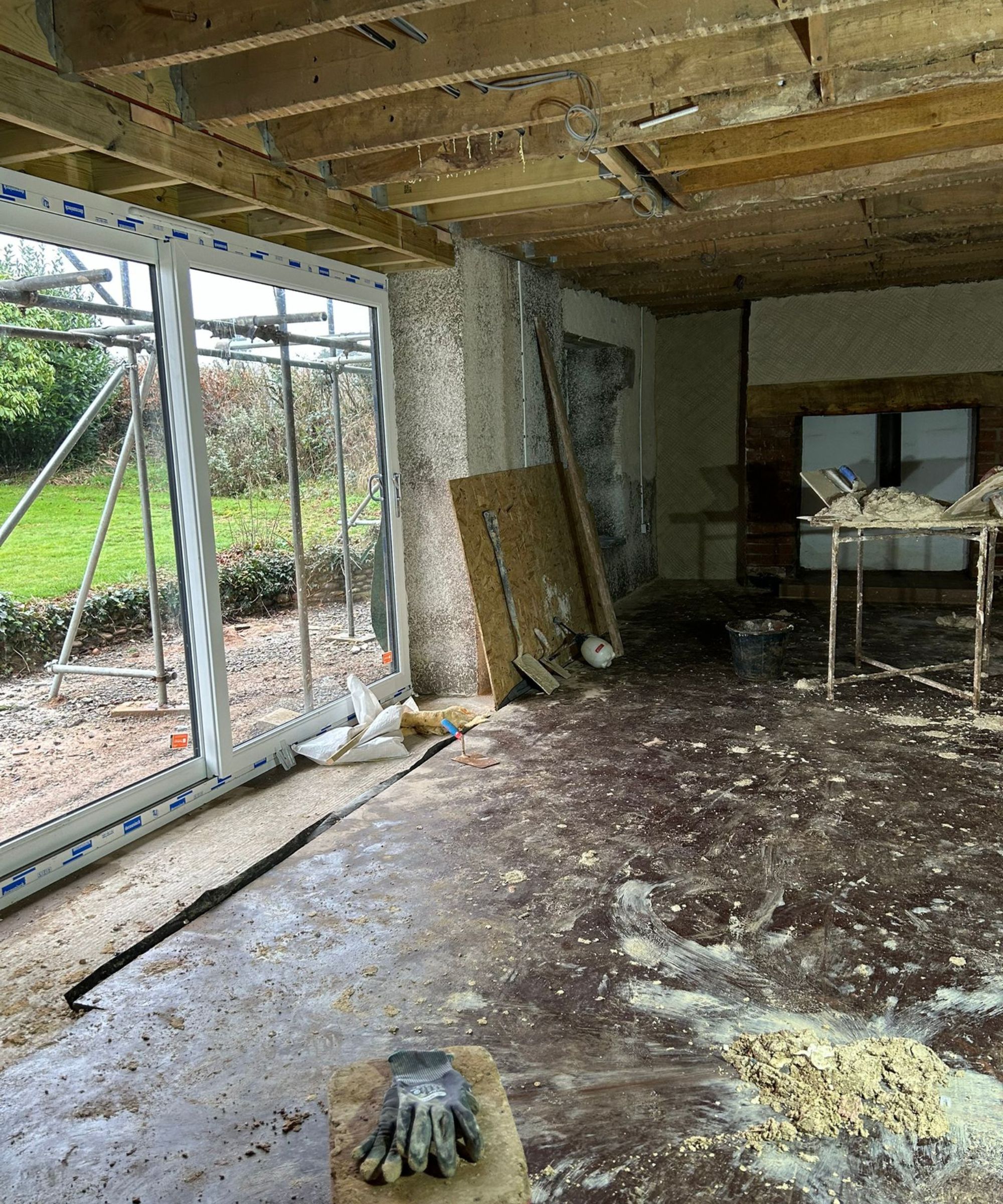 A large room under renovation with exposed ceiling joists, unfinished walls and construction materials scattered across the floor. A wide set of new sliding doors opens to a garden outside. Tools, scaffolding and debris create a busy work-in-progress scene.