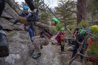 Melanie Brown, wearing one of her trademark leopard print jumpsuits, attempts to climb a mountain using ropes and a harness. She looks terrified. A climbing guide is beside her, while Ruby and the camera crew can be seen behind her