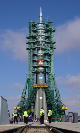 A green cage of support arms and structural access levels encase a green/grey soyuz rocket. People in white hard hats wearing blue stand at the base of the rocket. Six people closer to the foreground wearing yellow highlighter vests, but one is barely visible, as they are standing behind a small structure on the left.