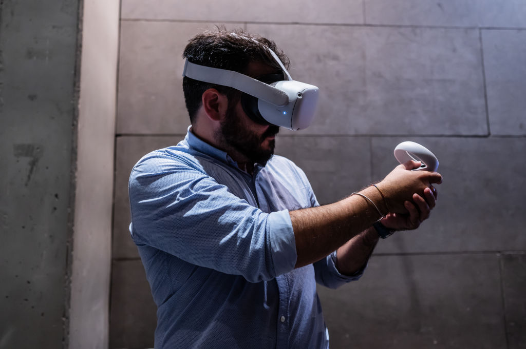 A man wearing a virtual reality headset, a blue button up shirt and two hand-held accessories looks to the left of the image