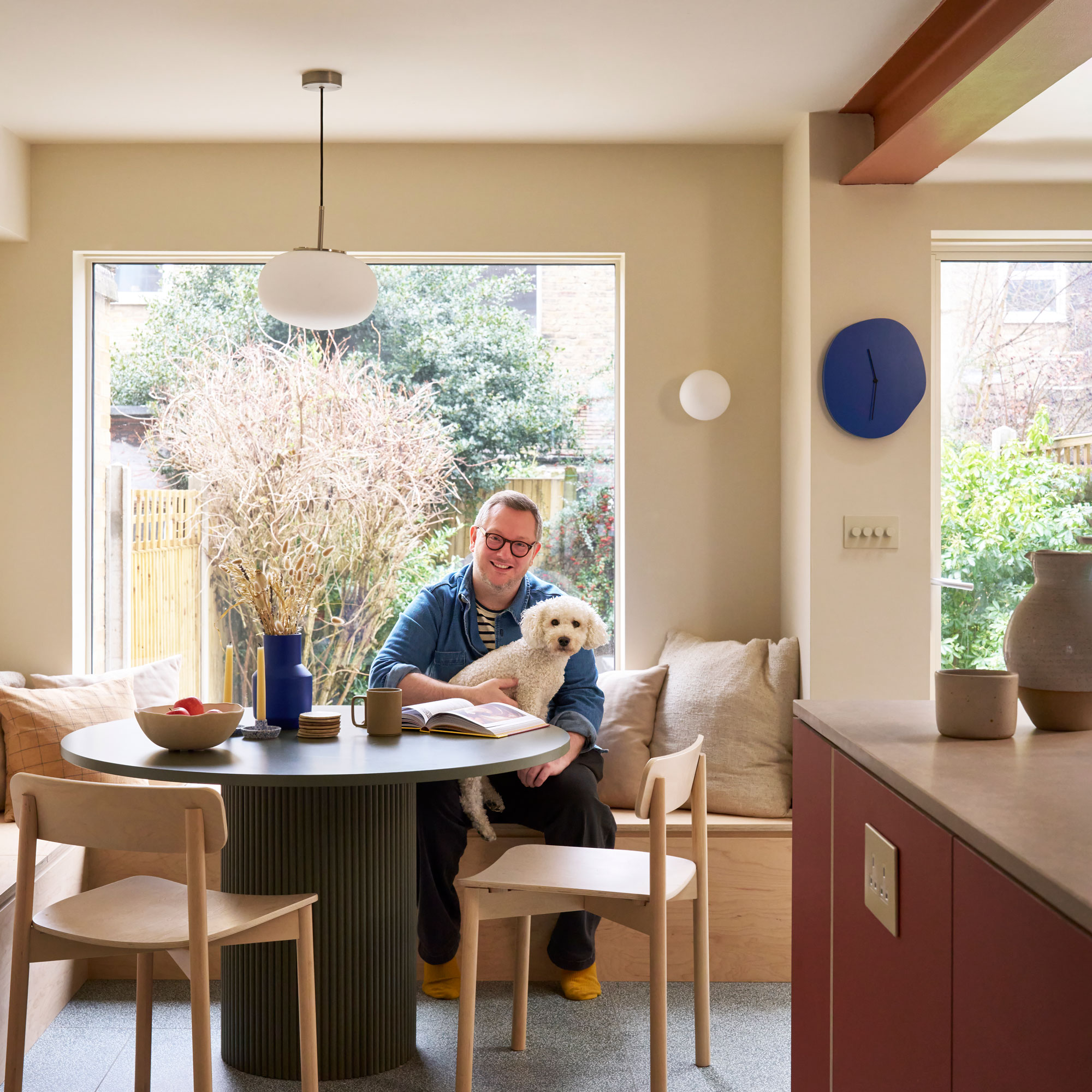A man holding a dog, sat at a round wooden table with a vase and picture window behind