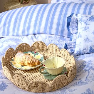 A bed dressed in a blue and white toile de jouy bed linen with a wicker scalloped tray holding a flower-shaped cup and a a side plate with croissant