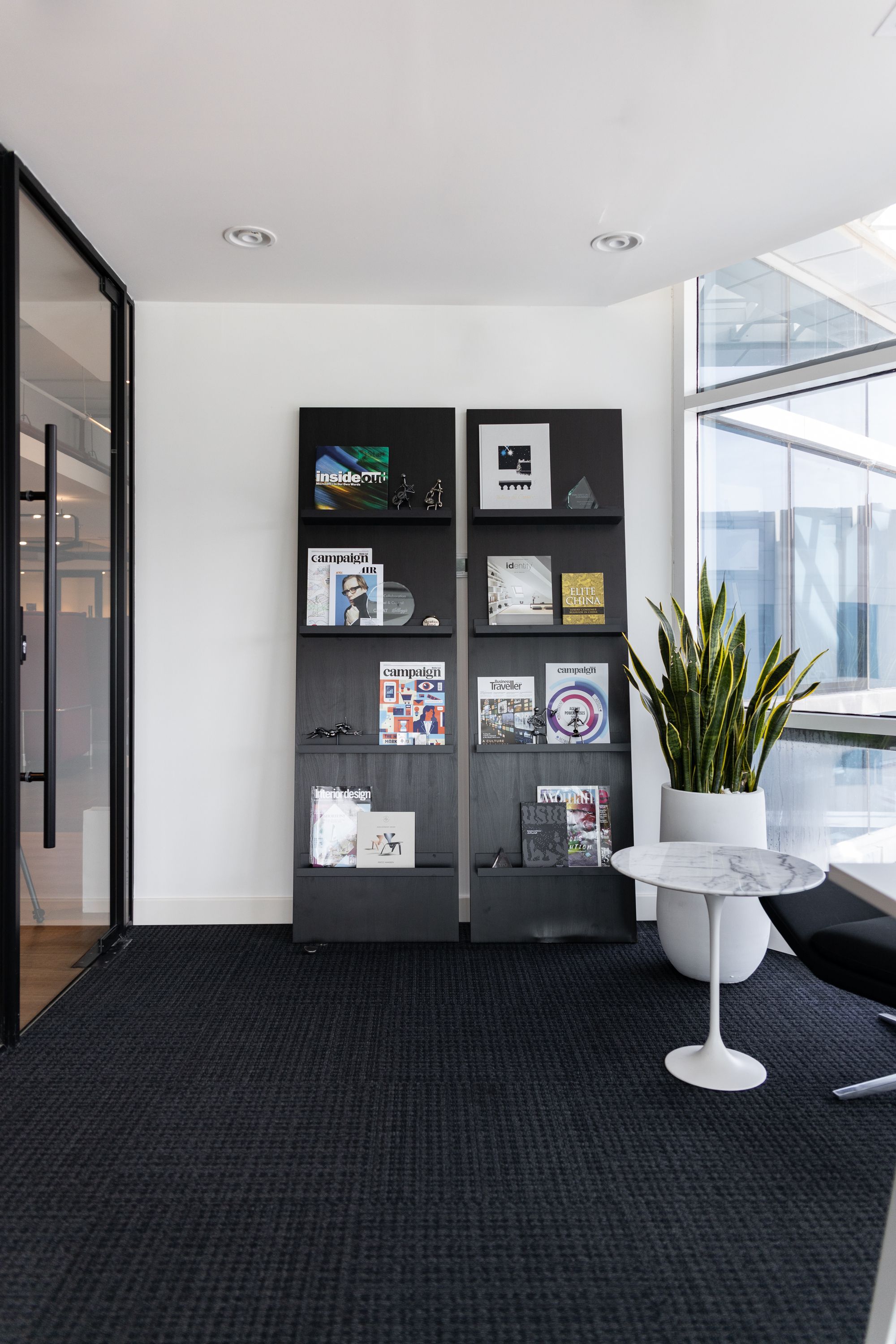 A white office room with black carpets and two large black magazine shelves leaning against the wall. There is also a white vase with a large green plant in it, a white marble table, and a black office chair.