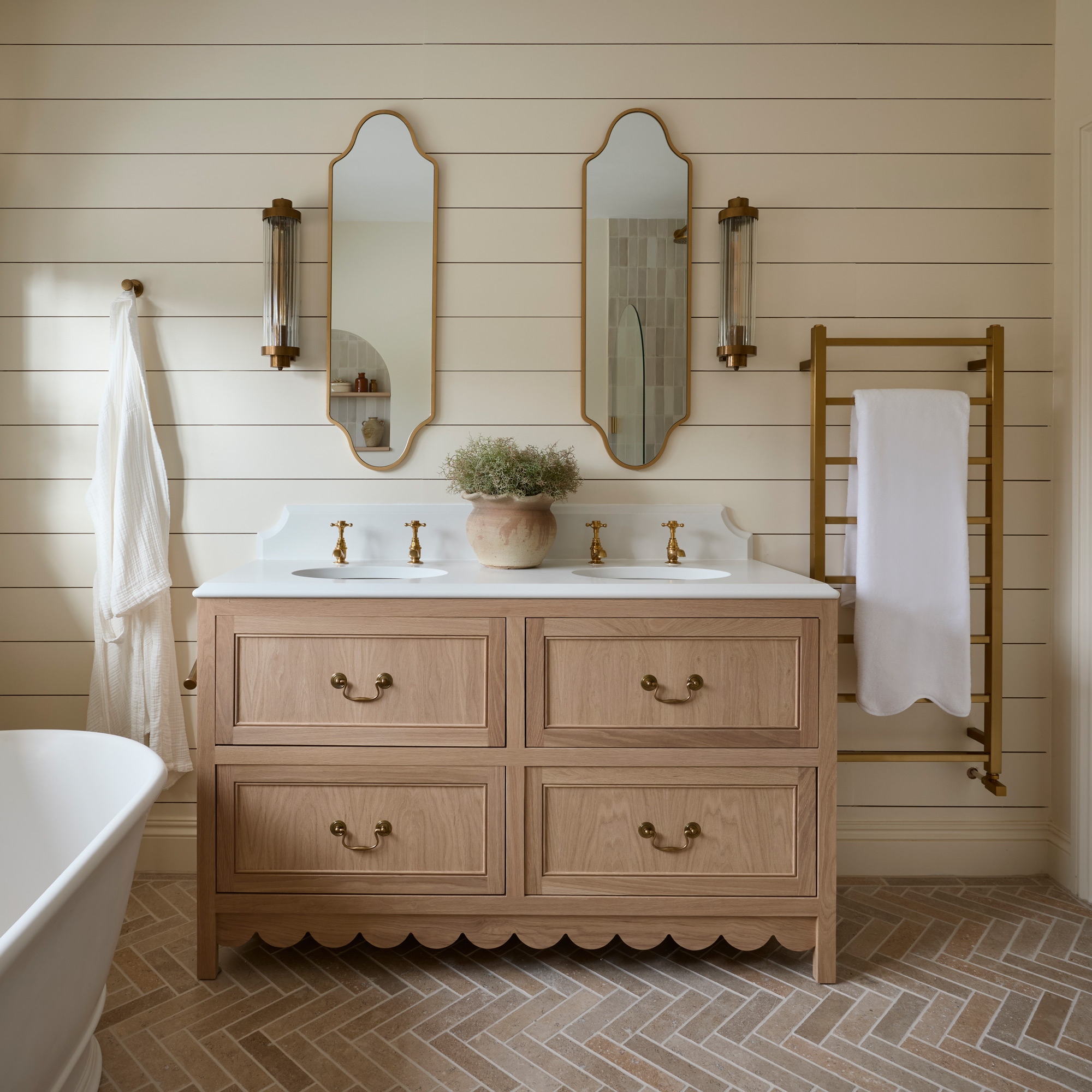 a neutral bathroom with horizontal wall panelling, a large white freestanding bath tub and brass fixtures, double vanity unit, and limestone herringbone floor tiles