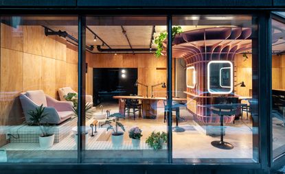 View through glass windows of the Le Hideout nail bar interior featuring wood covered walls, grey floors, pink and black chairs, manicure stations and green plants in white pots