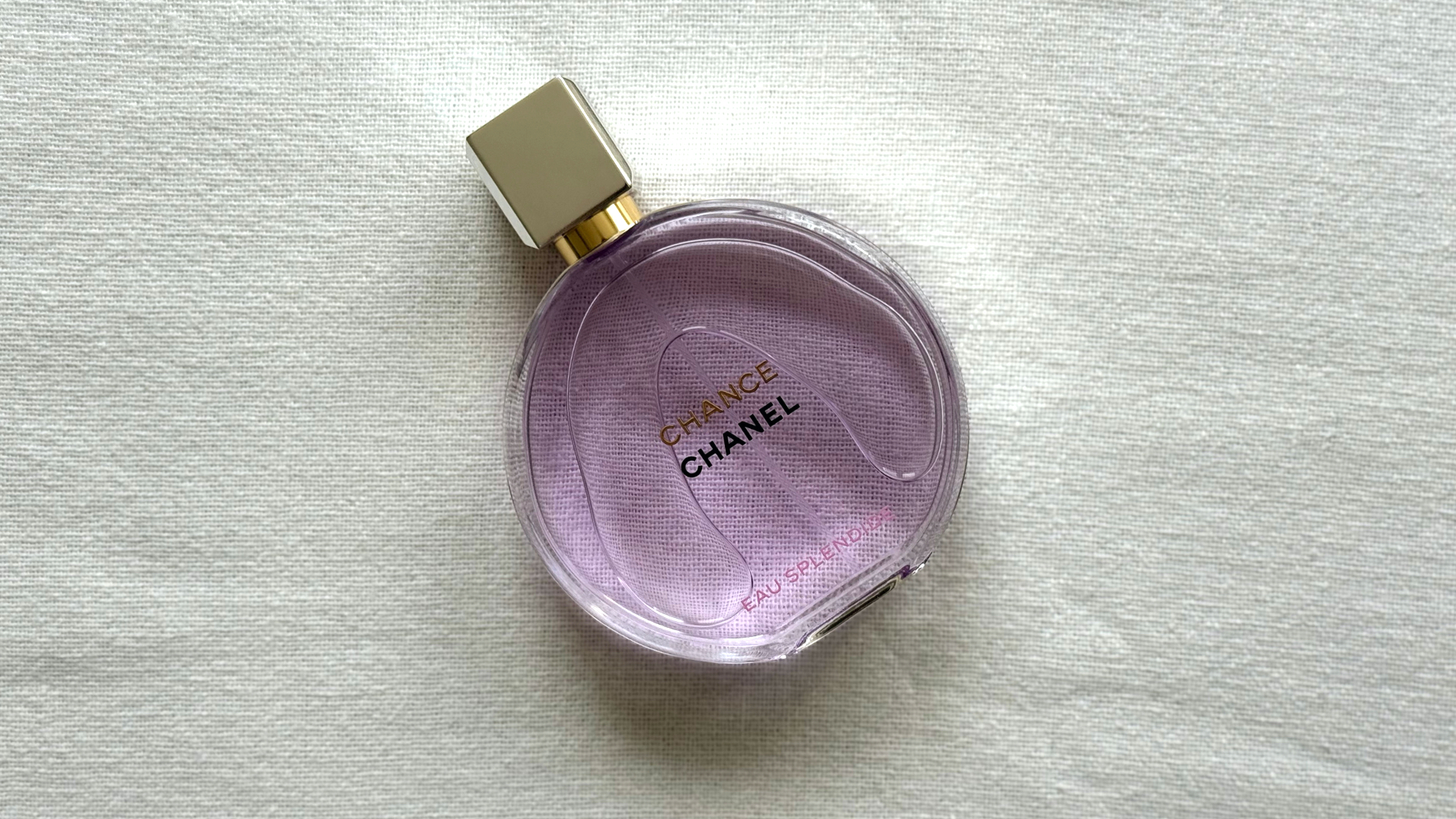 Bird's eye view of a bottle of Chanel Chance Eau Splendide, a violet perfume, laying at an an angle against an off-white linen background