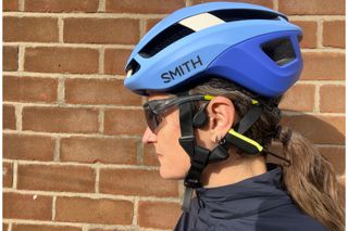 The Haylou PurFree Lite on a woman's head who is also wearing a blue helmet, sunglasses and standing side on in front of a brick wall