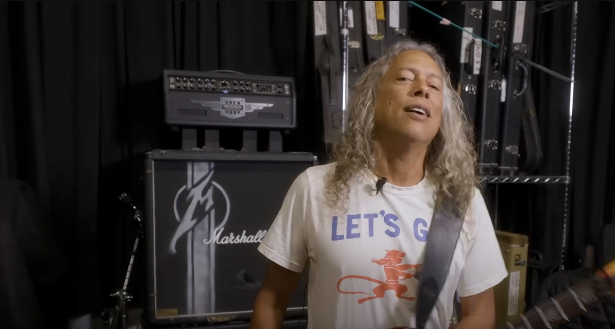 A still from a YouTube video featuring Kirk Hammett in a white T-shirt with &quot;Let&#039;s Go&quot; written on the front. Guitar amps are in the backgound.