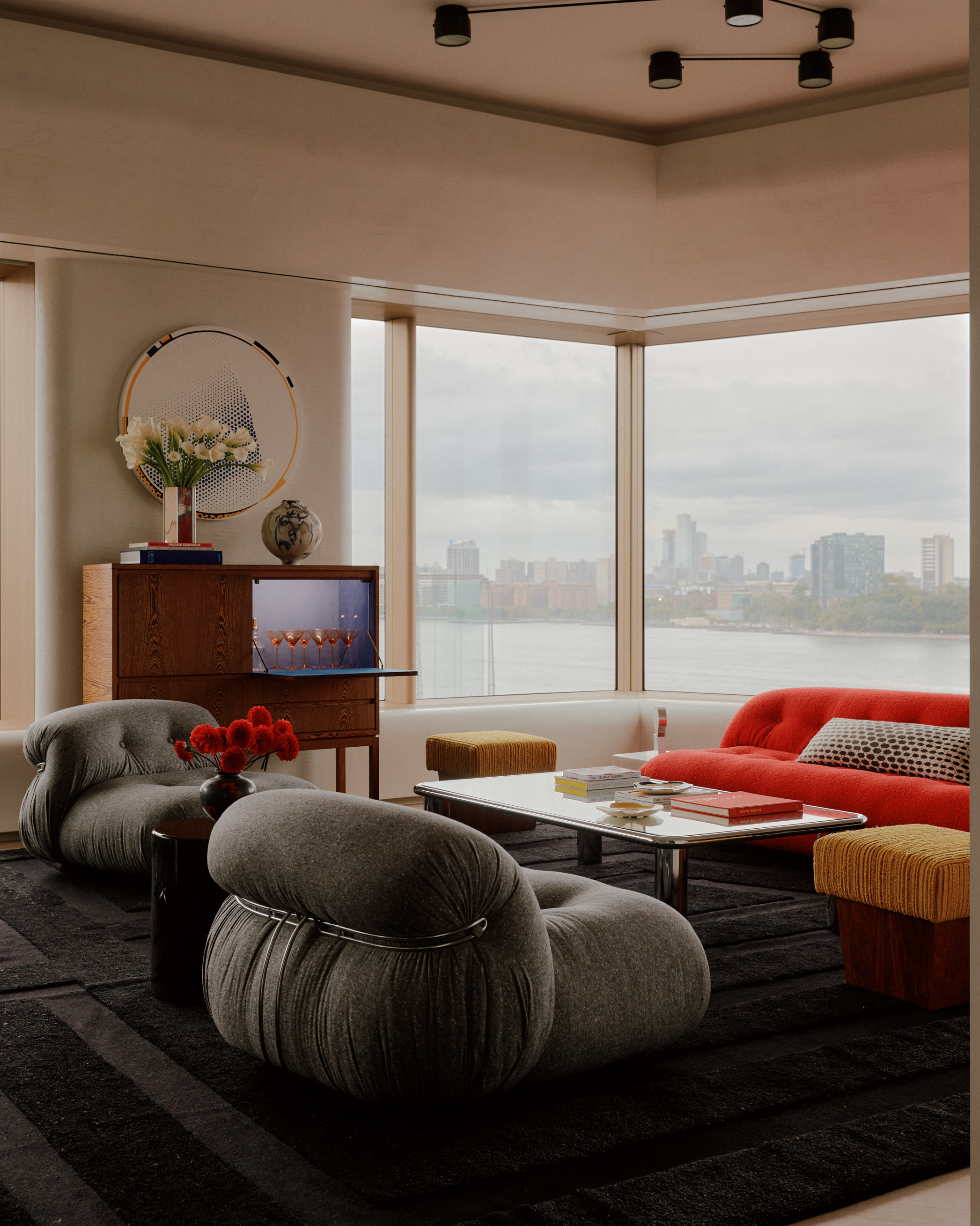 modern living room with gray armchairs, red sofa, metallic coffee table, bar cupboard with vase of flowers, and view over NYC